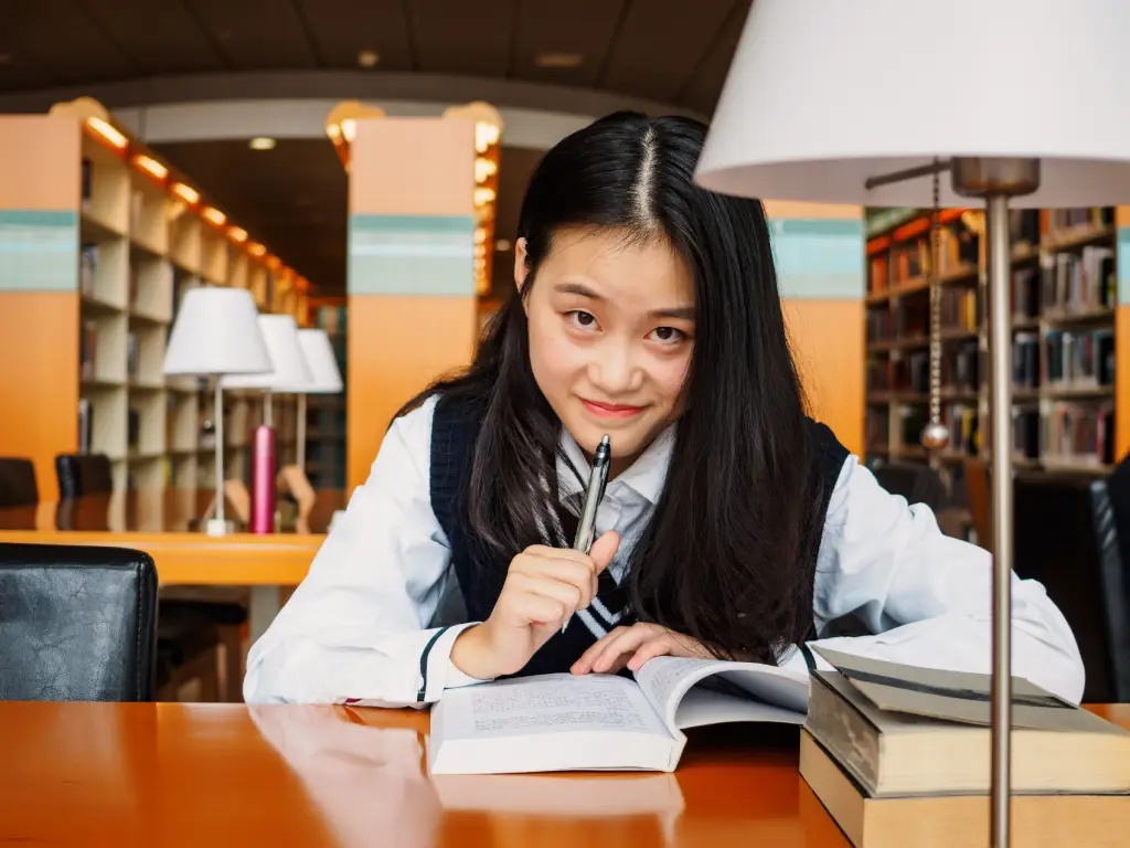 Chinese schoolgirl holds an open book at a library desk, looking at the camera with shelves behind her.