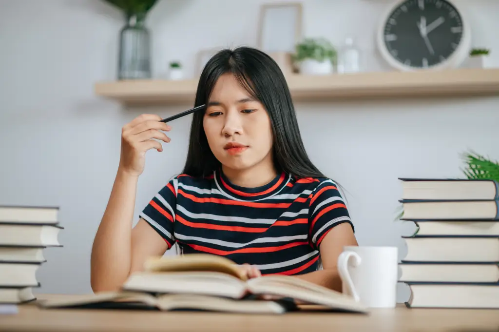 Teenage girl studying at a desk in a sunlit room, writing in a notebook with books and pencils nearby.