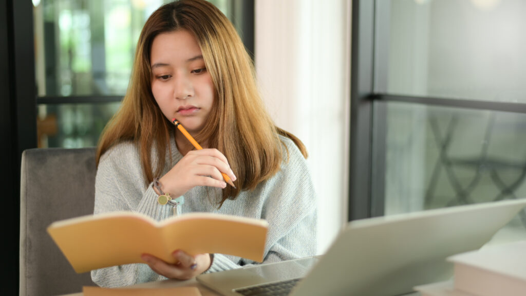 Young Asian student reviewing study notes beside a laptop during focused H2 Math revision and exam preparation.